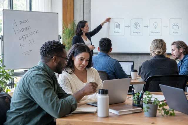 A hands-on AI workshop with a facilitator pointing at a simple workflow diagram while adult learners work in pairs on laptops.