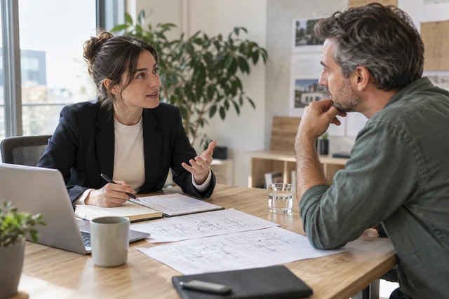 A business analyst interviewing a client with a paper checklist and product flow sketches spread on a table.